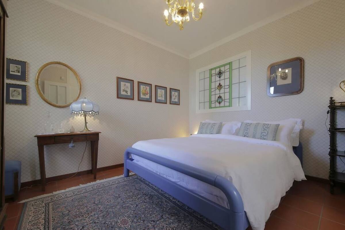 Ein Schlafzimmer mit Bett, Nachttisch, Lampe, Spiegel, gerahmten Bildern und einem Fenster mit Gitterstruktur.