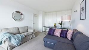 A modern living room with a grey sofa, a blue sofa, and a beige sofa. There is a coffee table with a lamp and a vase with flowers. A mirror is on the wall, and a door is visible in the background. - Deluxe Kensington Apartment With Balcony (London)