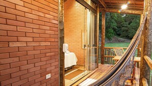 Un hamac sur une terrasse en bois, avec un mur en briques et une porte vitrée donnant sur une pièce où se trouve un lit.