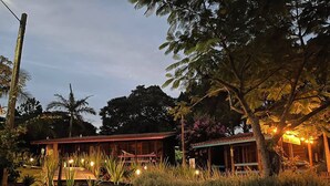 Un bâtiment en bois à deux étages, surmonté d’un toit rouge, entouré d’une végétation luxuriante et d’herbes hautes.
