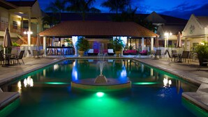 A swimming pool with a fountain, surrounded by lounge chairs and tables, with a covered area and palm trees in the background. - Hotel North Resort (Paramaribo)
