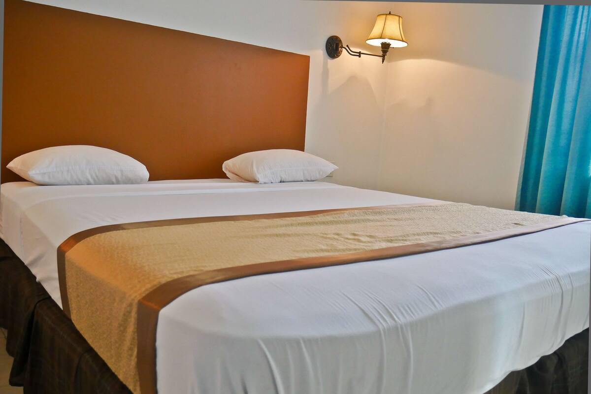 Una cama doble con ropa blanca, un plaid color beige y una lámpara de pared.