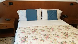 Hotellihuone, jossa on puinen pääty, kukkakuviollinen päiväpeite, kaksi sinistä tyynyä, yöpöytä, jossa on kaukosäädin, ja puhelin yöpöydällä.