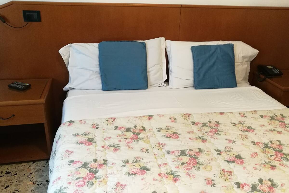 A hotel room with a wooden headboard, a floral bedspread, two blue pillows, a bedside table with a remote control, and a phone on the nightstand.