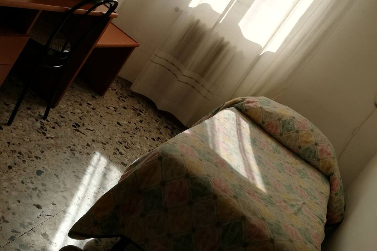 A room with a bed, a desk with a computer, a chair, a window with curtains, and a wall-mounted picture.