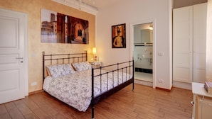 A bedroom with a bed, a chandelier, a painting, a bathroom door, and a white wardrobe. - Cento Passi Dal Duomo (Arezzo)