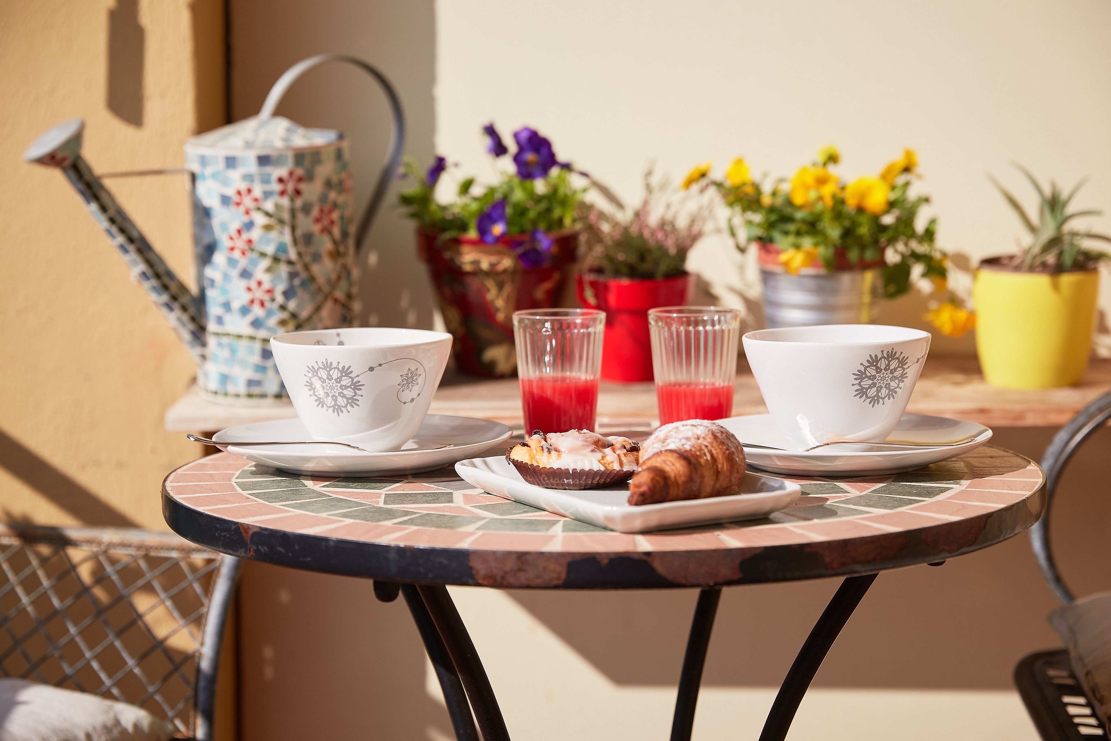 Une table ronde avec un plateau carrelé, deux tasses blanches ornées de motifs floraux, deux verres transparents contenant un liquide rouge, un croissant et une petite assiette avec une pâtisserie.