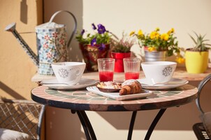 Una mesa redonda con cubierta de azulejos, dos tazas blancas con motivos florales, dos vasos transparentes con líquido rojo, un croissant y un plato pequeño con un pastel.
