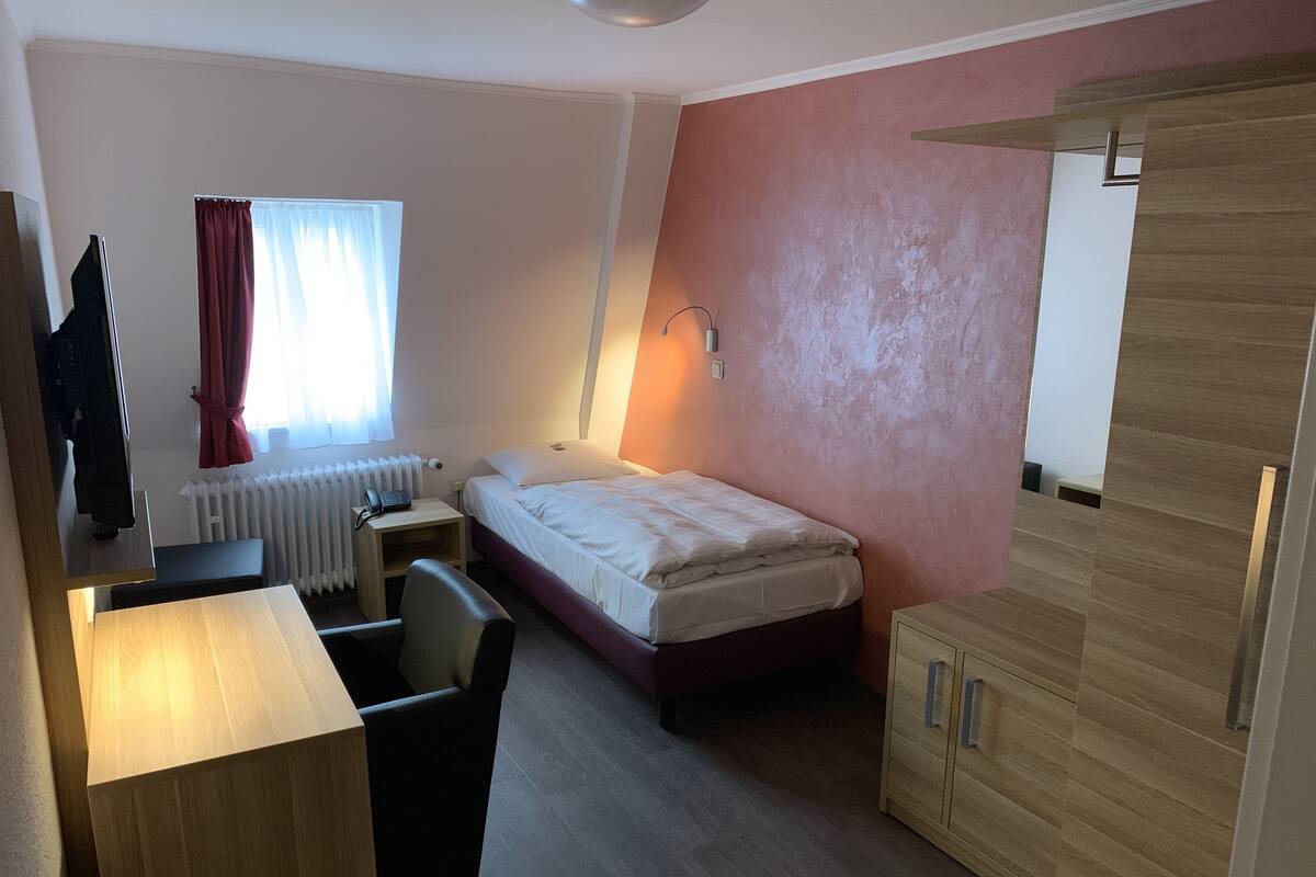 A hotel room with a bed, a desk, a chair, a radiator, and a wardrobe.