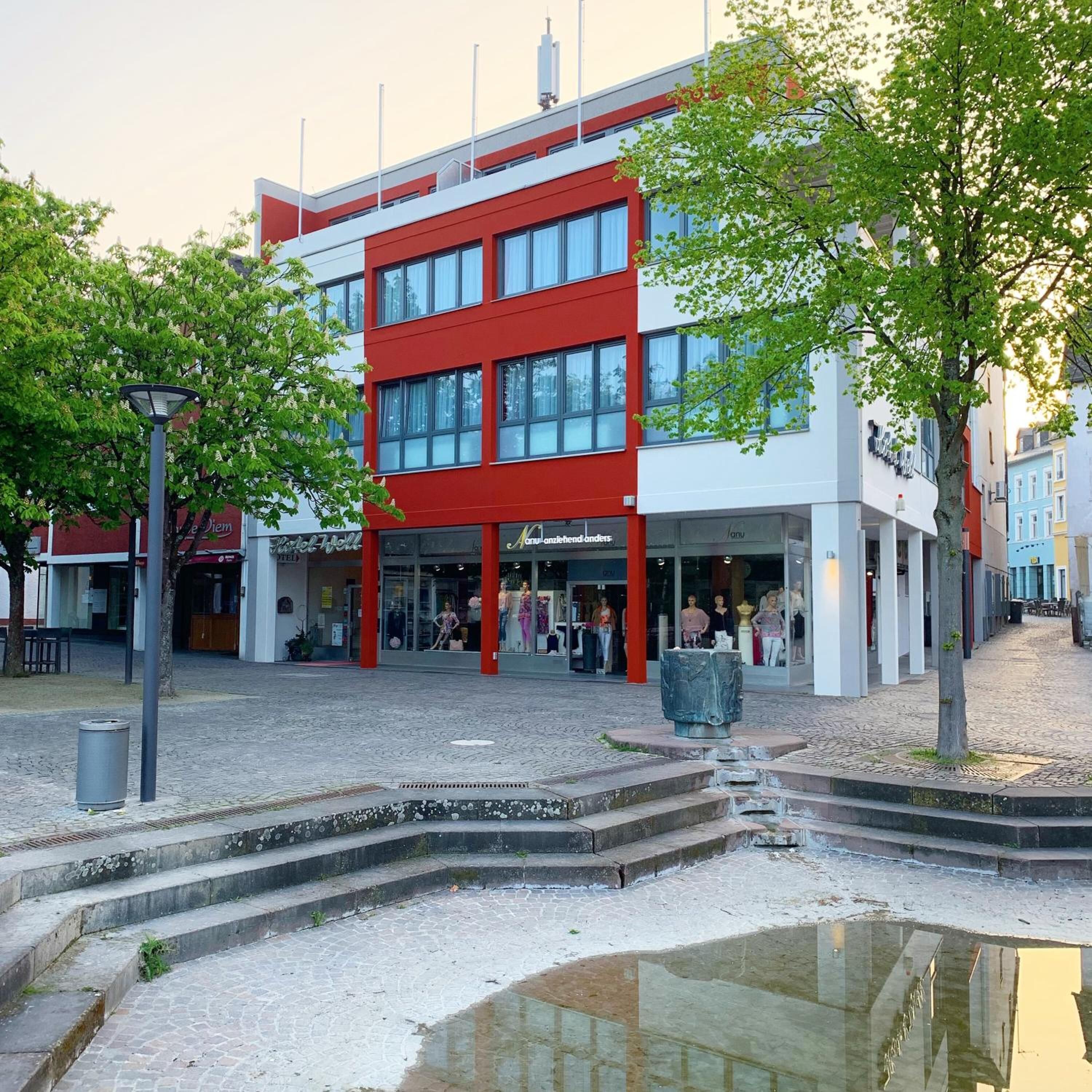 Un bâtiment moderne à la façade rouge et blanche, doté d’une entrée centrale et d’un petit espace extérieur avec des marches menant à une fontaine.