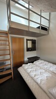 Dormitorio con literas, tragaluz, escalera de madera, puerta de madera, estante negro y colchón blanco.