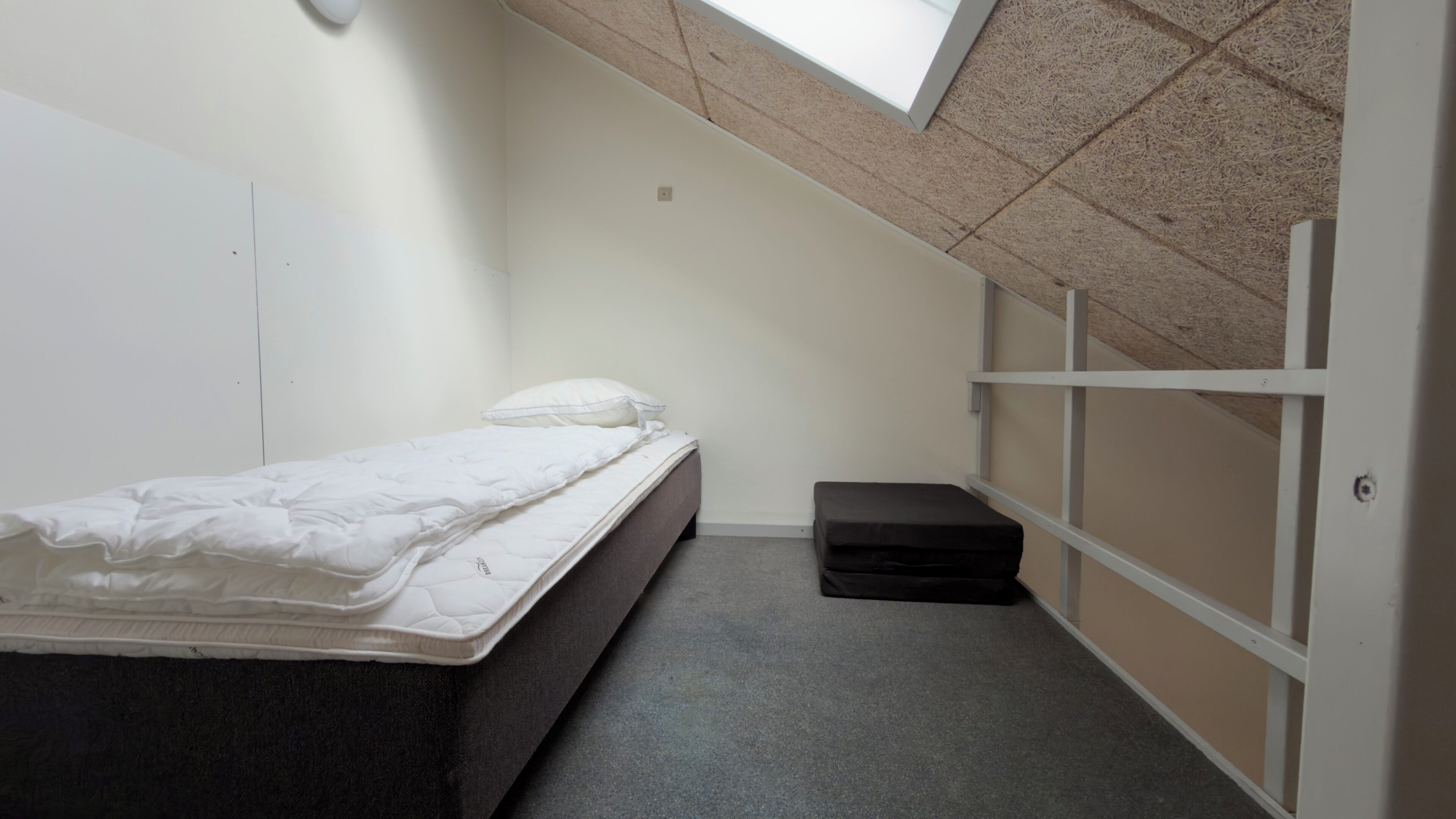 Een kleine kamer met een enkel bed, schuine wand, een bankje en uitzicht op de lucht.