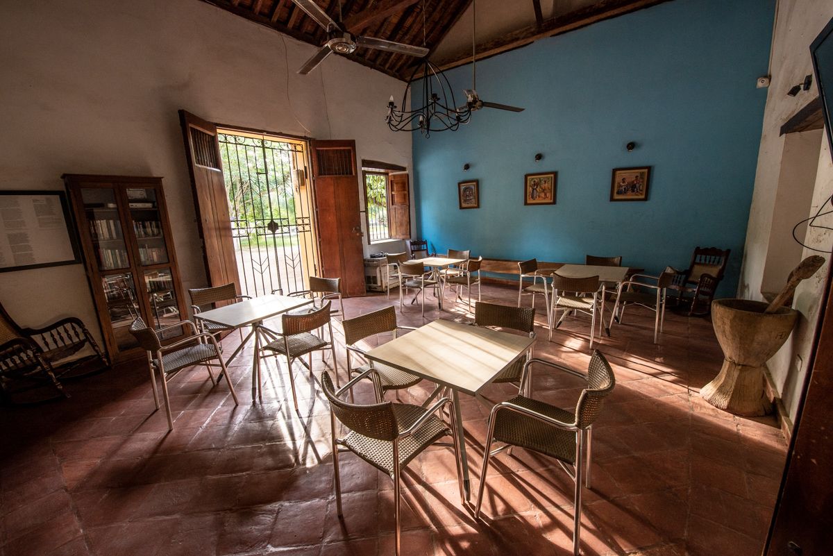 A spacious room with a high ceiling, a ceiling fan, and a large window allowing natural light. There are multiple tables and chairs arranged for dining or meetings.