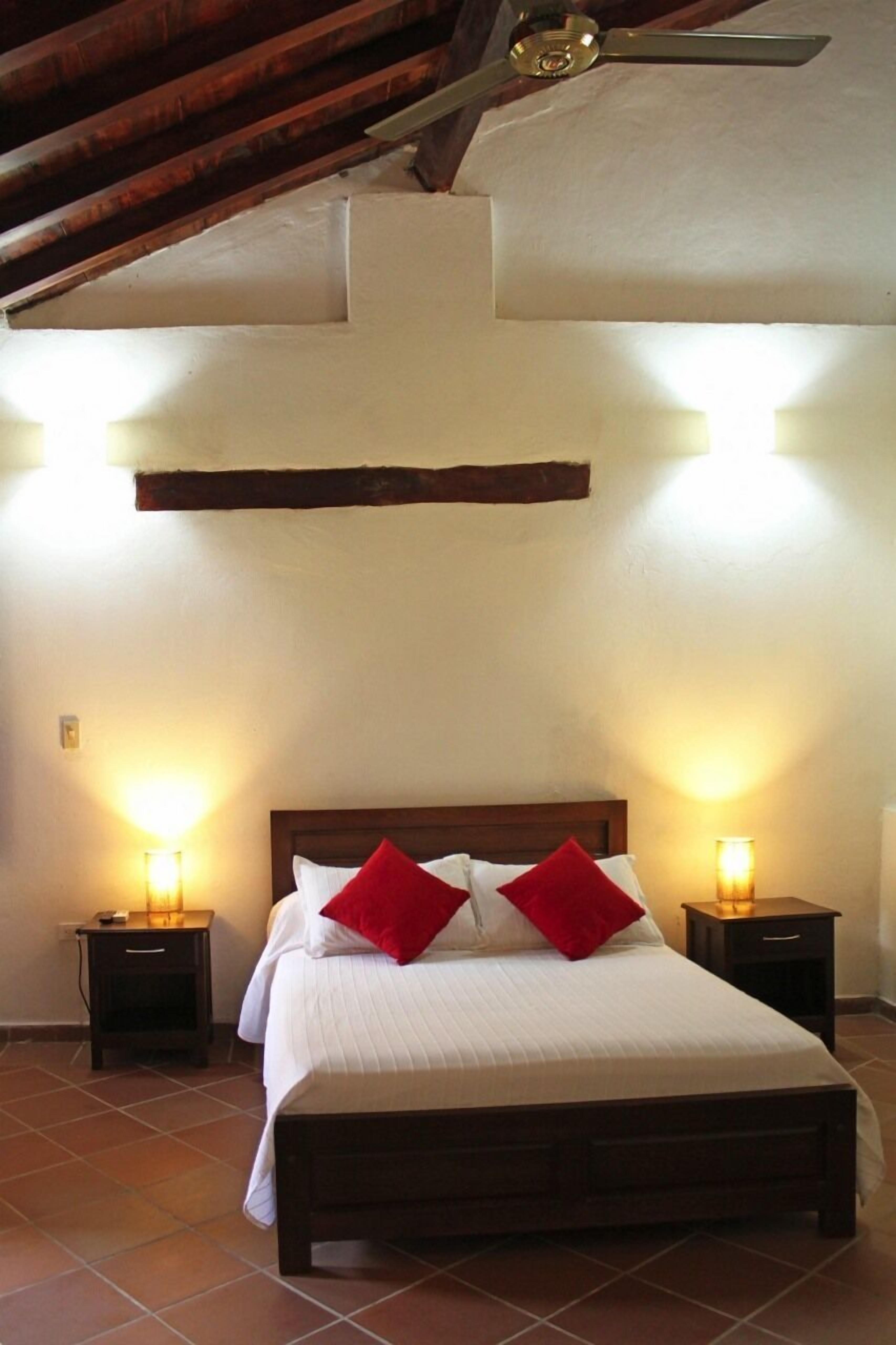 Un dormitorio con una cama de madera, dos mesitas de noche con lámparas y un techo inclinado con vigas a la vista.