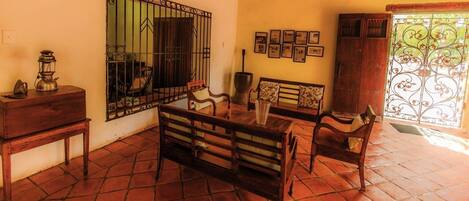 Una habitación con suelo de baldosas, una mesa de madera con sillas, un armario de madera, un espejo con marco de metal y una ventana con reja decorativa.