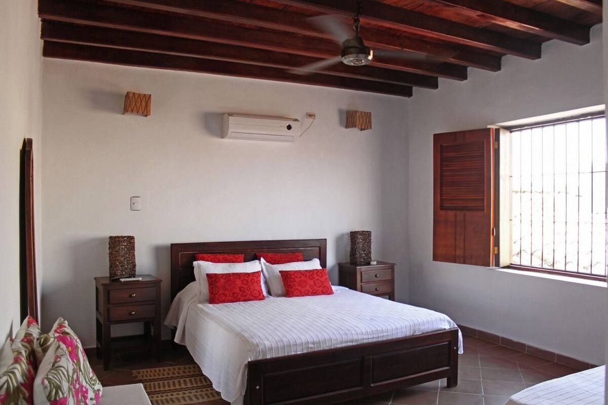 A bedroom with a wooden ceiling, a bed with red and white bedding, a bedside table with a lamp, and a window with bars.