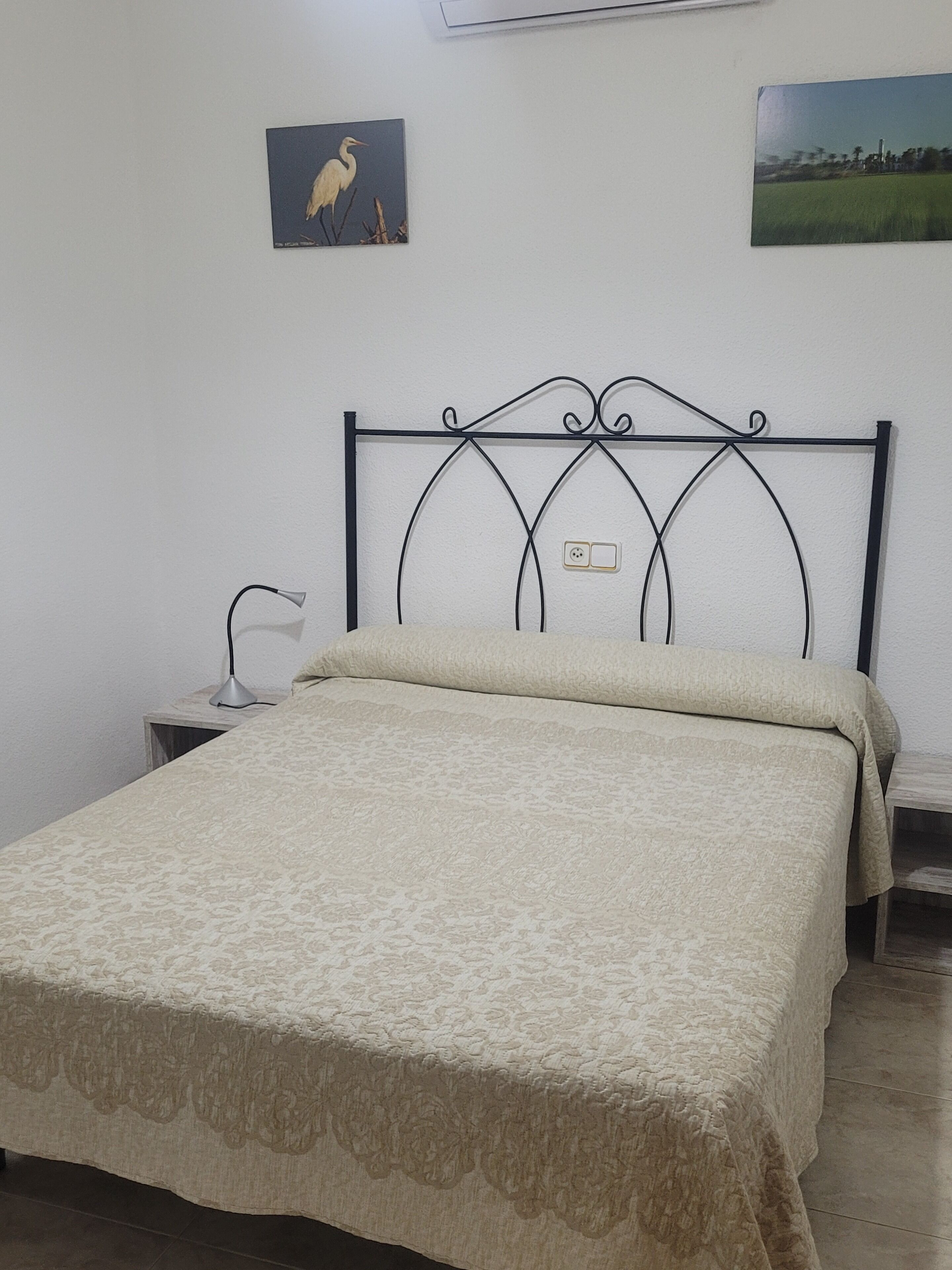 Een bed met een metalen hoofdbord, een nachtkastje met een lamp, en twee ingelijste foto’s aan de muur.