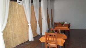 Um restaurante com divisórias de bambu, cortinas brancas e toalhas de mesa laranja.
