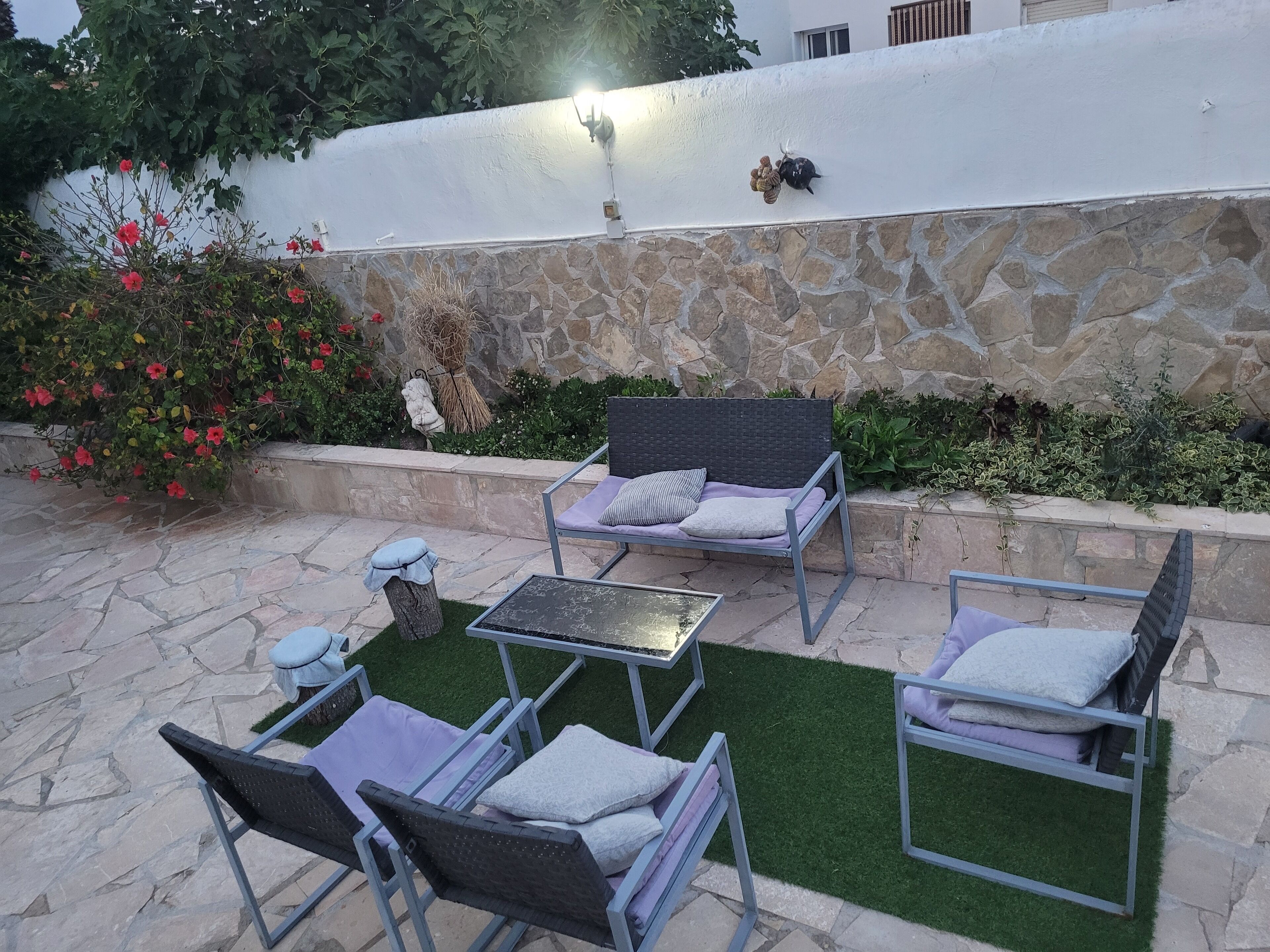 Un patio al aire libre con piso de piedra, pared de piedra y cerca blanca. Hay dos sillas de mimbre con cojines y una pequeña mesa.
