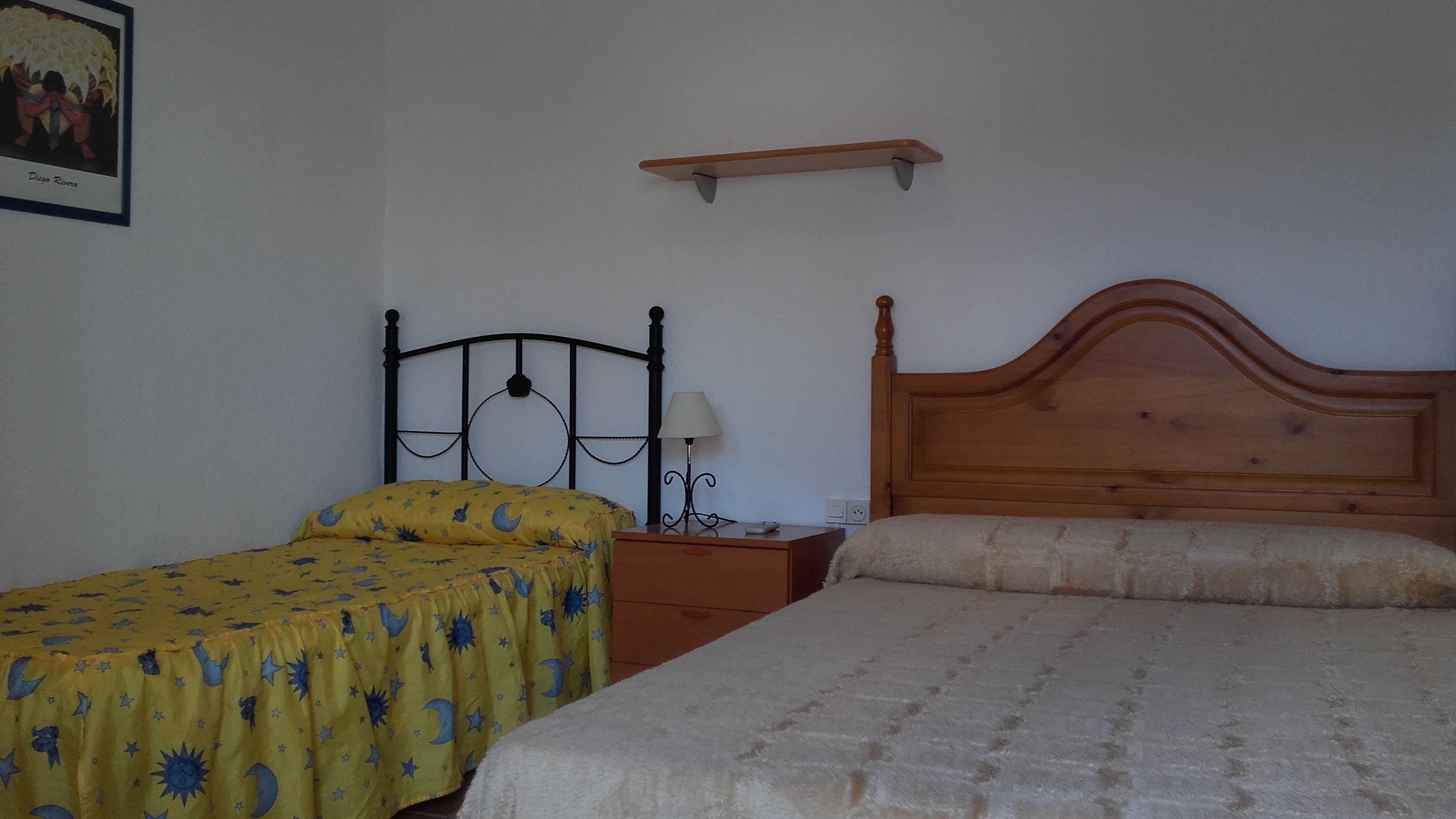 A bedroom with two beds, one with a yellow bedspread and the other with a white bedspread. There is a nightstand with a lamp and a small shelf above the bed.