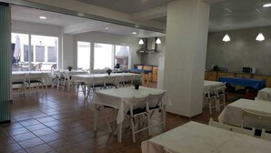 A spacious dining area with multiple tables and chairs, a kitchen in the background, and large windows allowing natural light. - La Llar de Laura (Amposta)