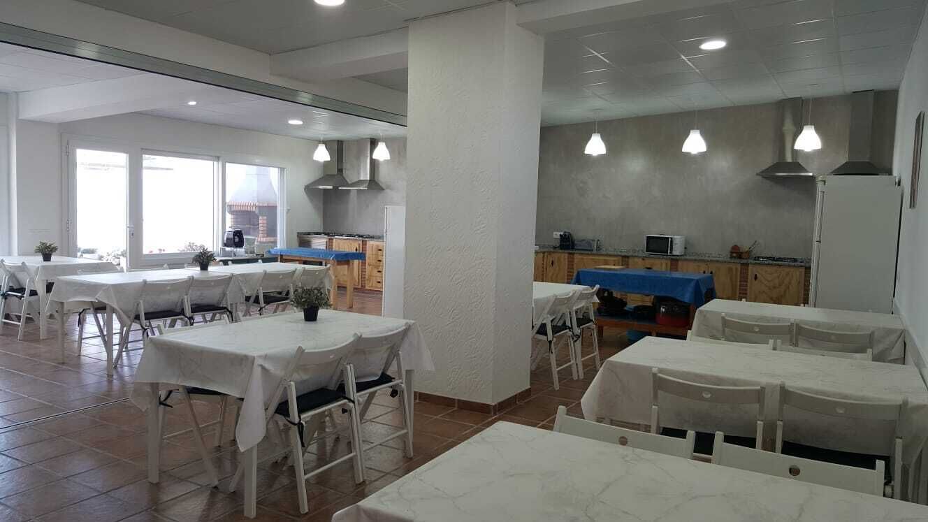 A dining area with white tables and chairs, a kitchen in the background, and large windows allowing natural light.