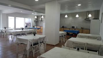 A dining area with white tables and chairs, a kitchen in the background, and large windows allowing natural light.