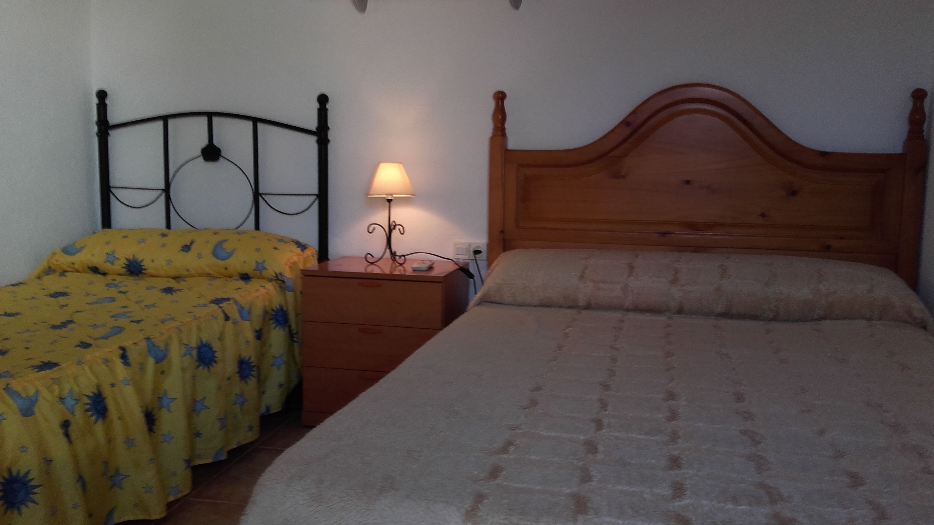 A bedroom with two beds, one with a yellow bedspread and the other with a beige one. There is a wooden nightstand with a lamp between the beds.