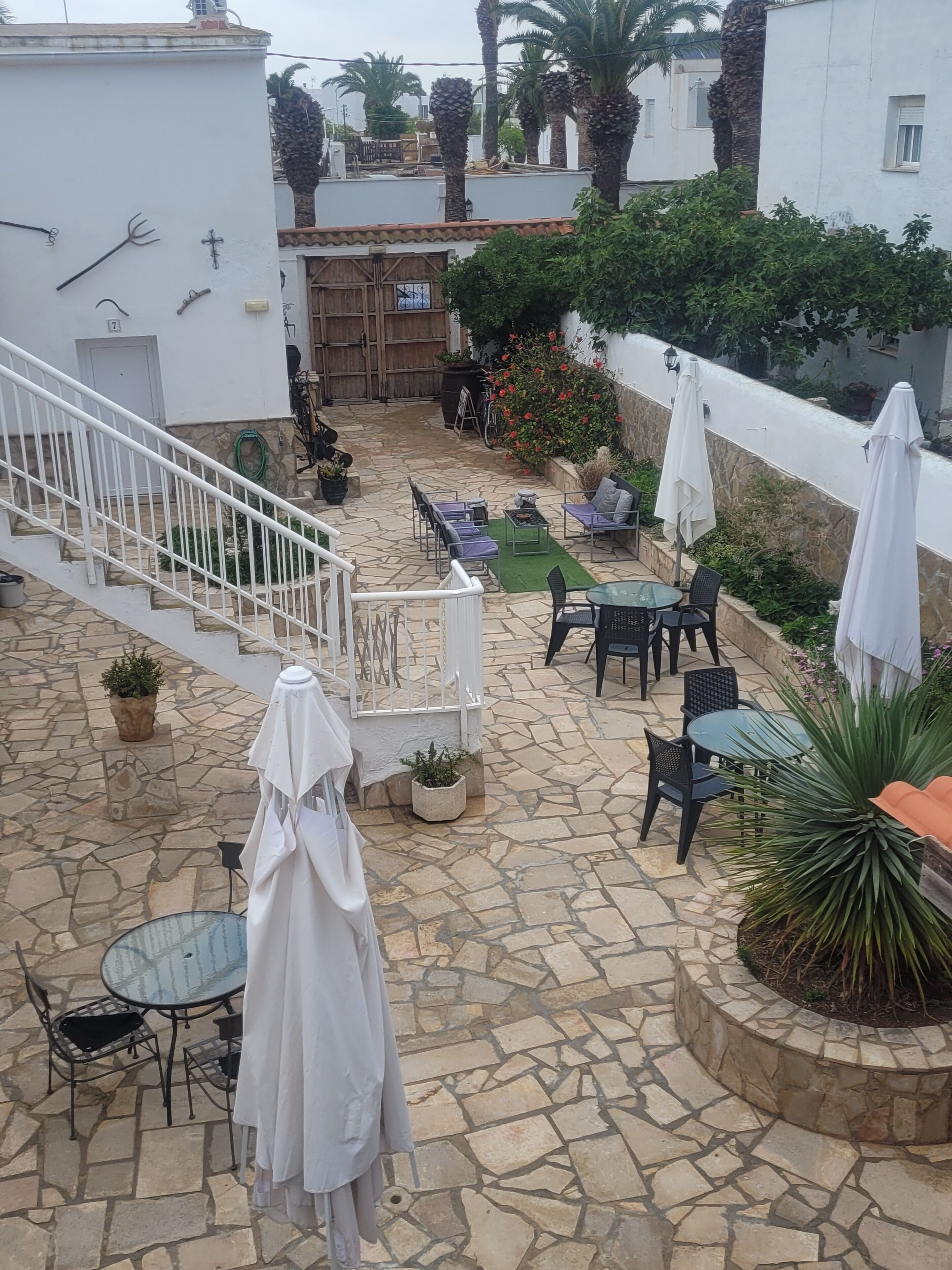 En terrasse med kampesten og udendørs møbler, herunder borde og stole, omgivet af krukker med planter og parasoller.