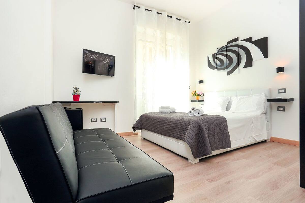 A modern bedroom with a bed, a black sofa, a small table with a red vase, and a wall-mounted TV.