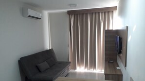 A modern living room with a grey sofa, a wooden coffee table, a flat-screen TV on a wooden stand, and a large window with curtains. - Atrium - Achei Ferias (Caldas Novas)