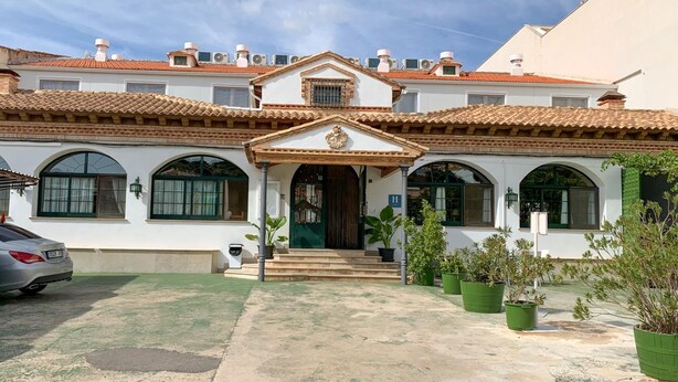 A two-story building with a tiled roof, a central entrance with a decorative arch, and a small porch. There are potted plants in front of the building.