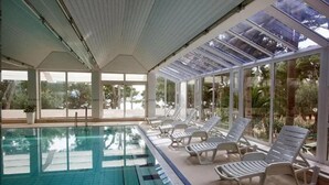 An indoor swimming pool with clear glass walls, white lounge chairs, and a tiled floor. - Biokovka (Makarska)