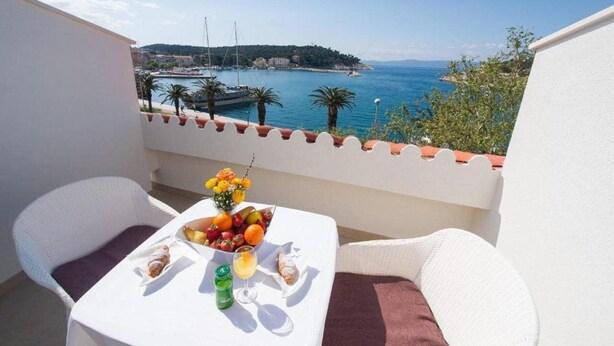 A balcony with white wicker furniture, a table with a fruit bowl, a bottle, and a glass. The view includes a body of water and a distant shoreline with buildings. - Biokovka (Makarska)