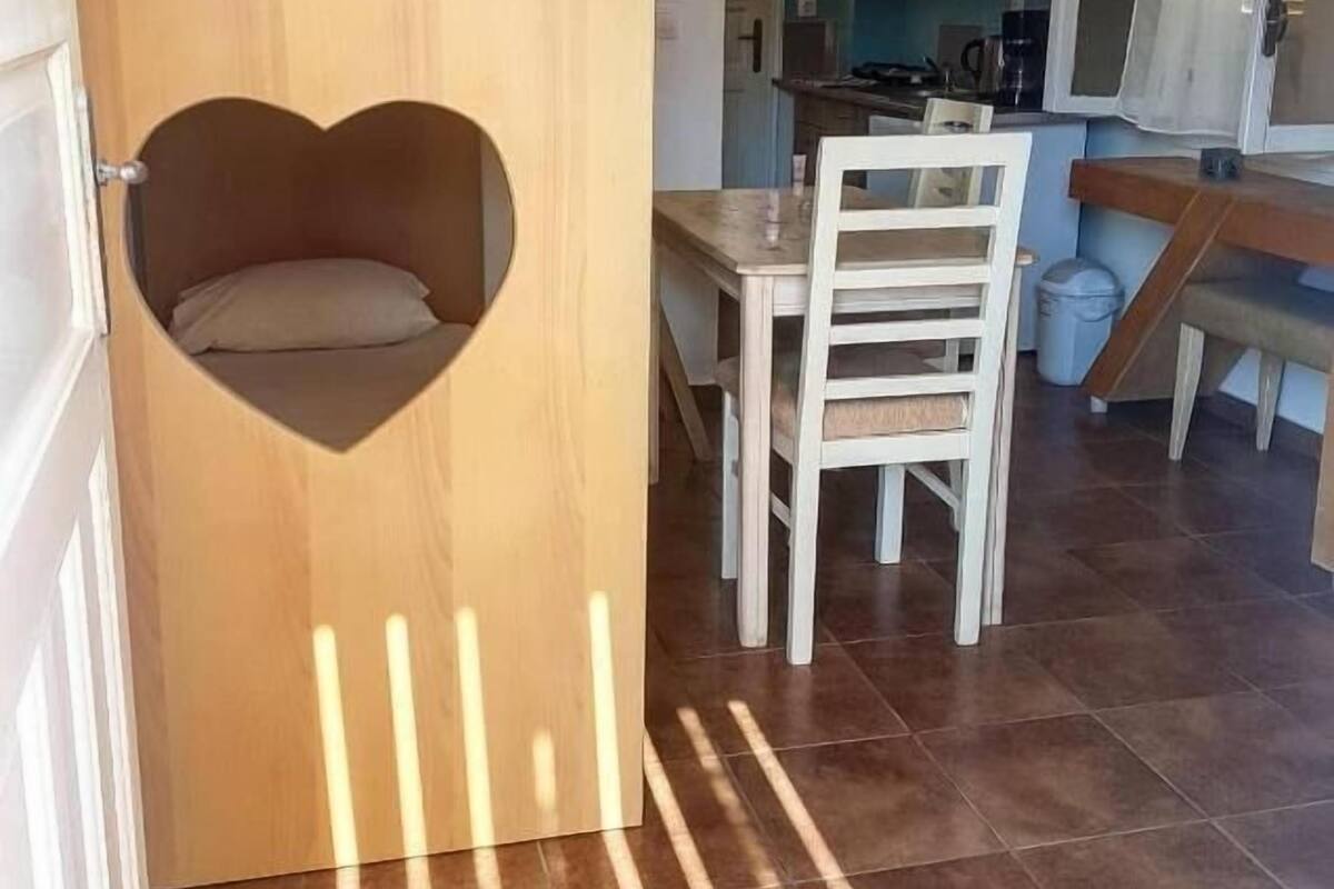 A room with a heart-shaped cutout in a wooden door, a dining table with chairs, and a kitchen area in the background.