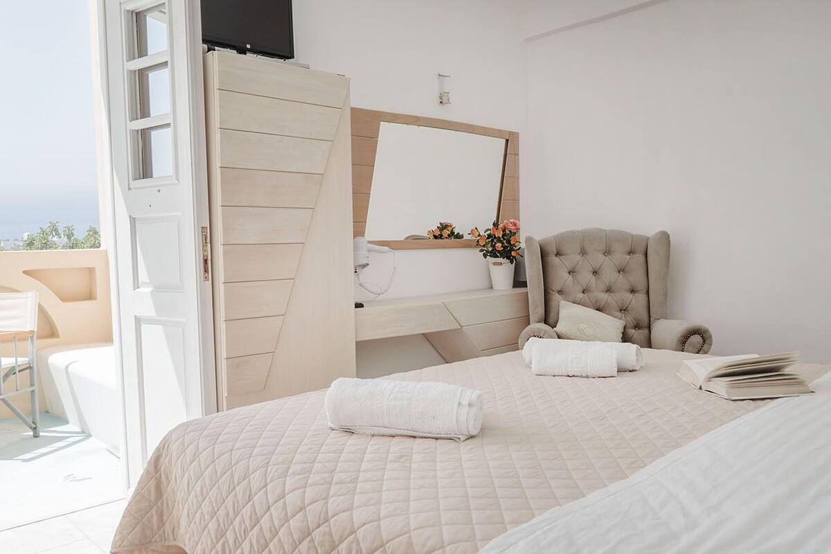 A bedroom with a bed, a chair, a vanity with a mirror, and a view of the outdoors.