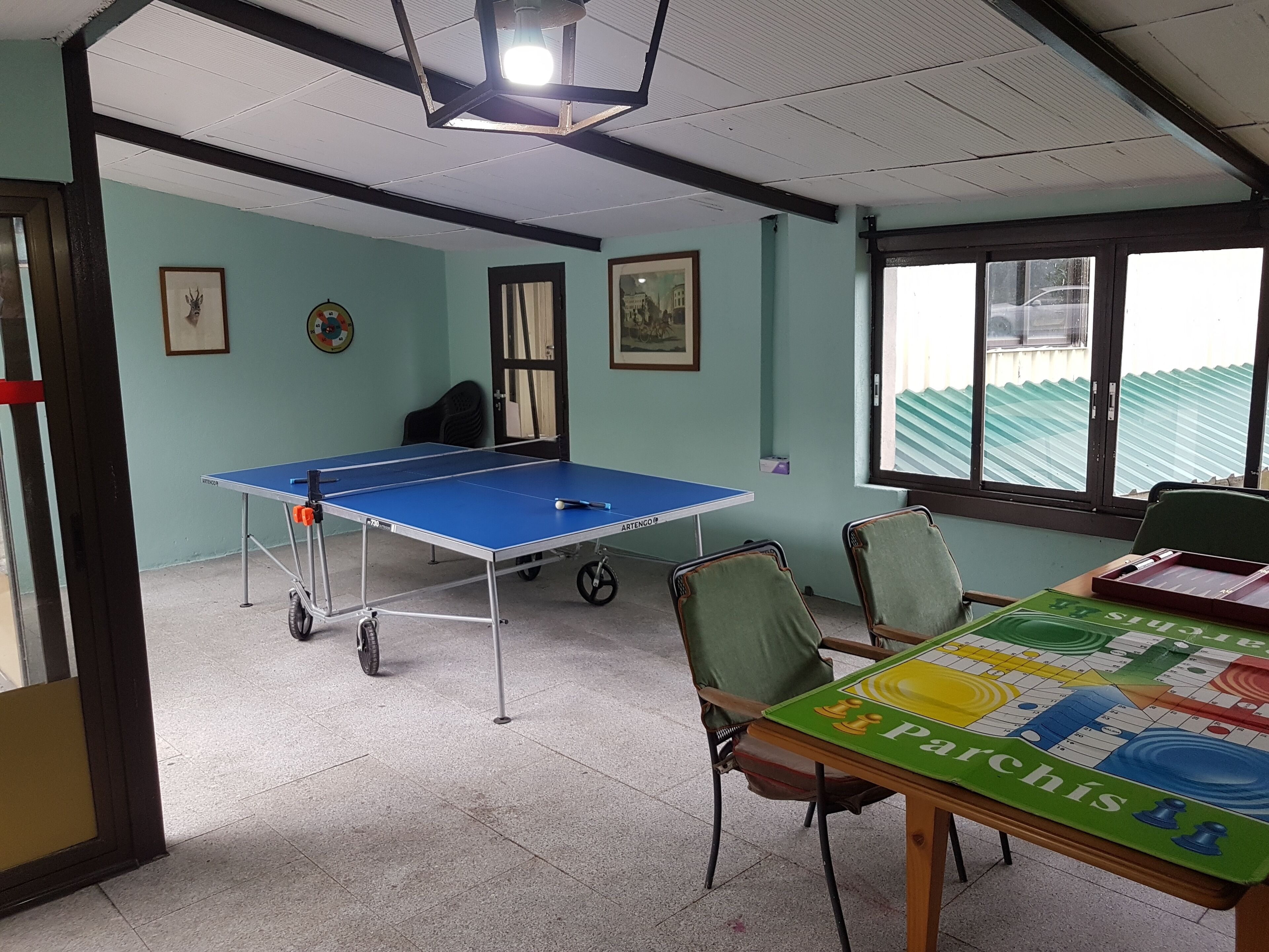Una habitación con mesa de ping pong, sillas, mesa de ajedrez y ventana con vista a un edificio.