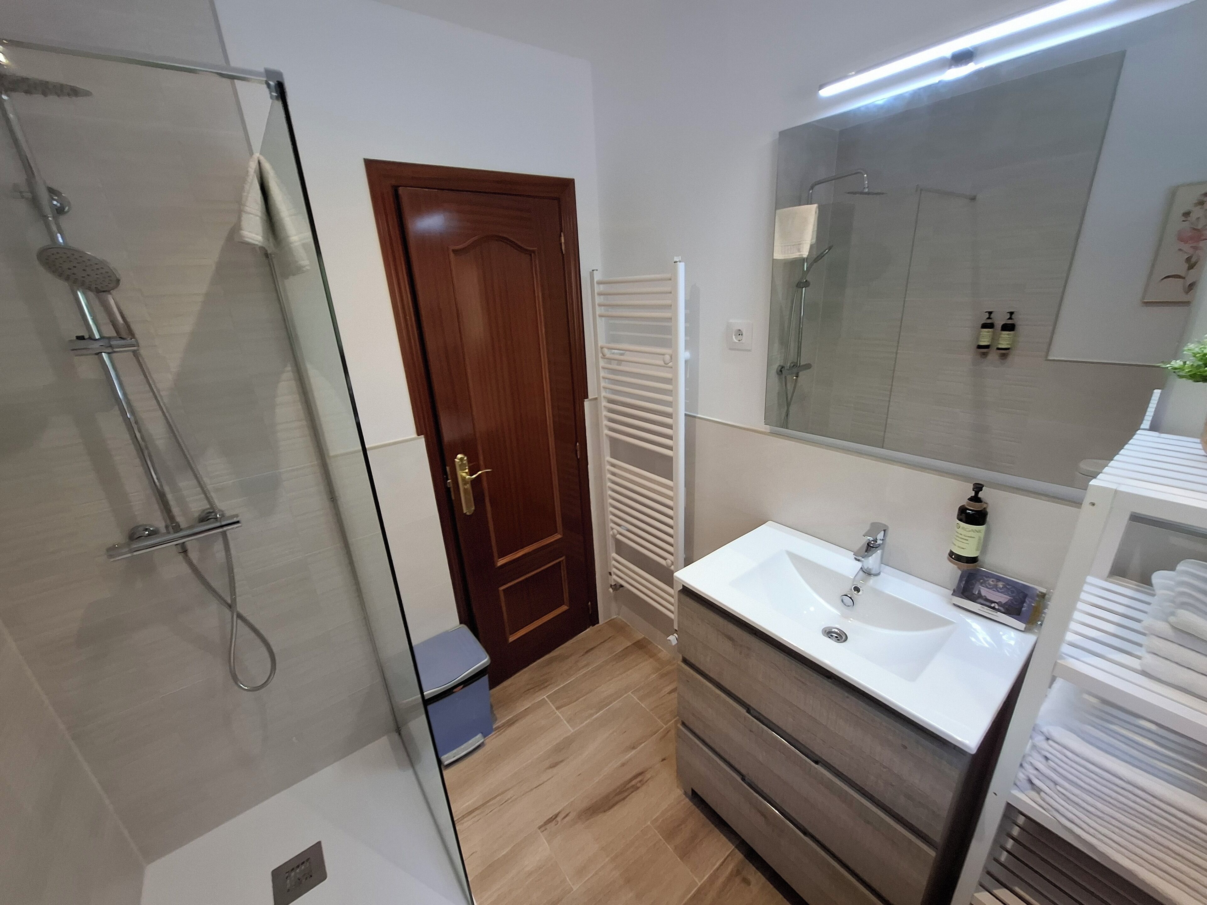 Une salle de bain moderne avec une douche à parois de verre, une porte en bois, un lavabo blanc avec un miroir au-dessus et un porte-serviettes.