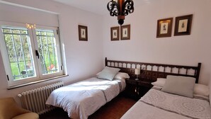 A bedroom with two single beds, a wooden floor, a radiator, a chair, and a window with a view of greenery.
