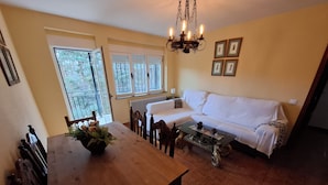 A dining area with a wooden table, chairs, and a chandelier. There is a white sofa and a window with blinds.