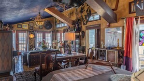 A room with a large bed, a dining table, and a chandelier. There are multiple windows and a ceiling with exposed beams.