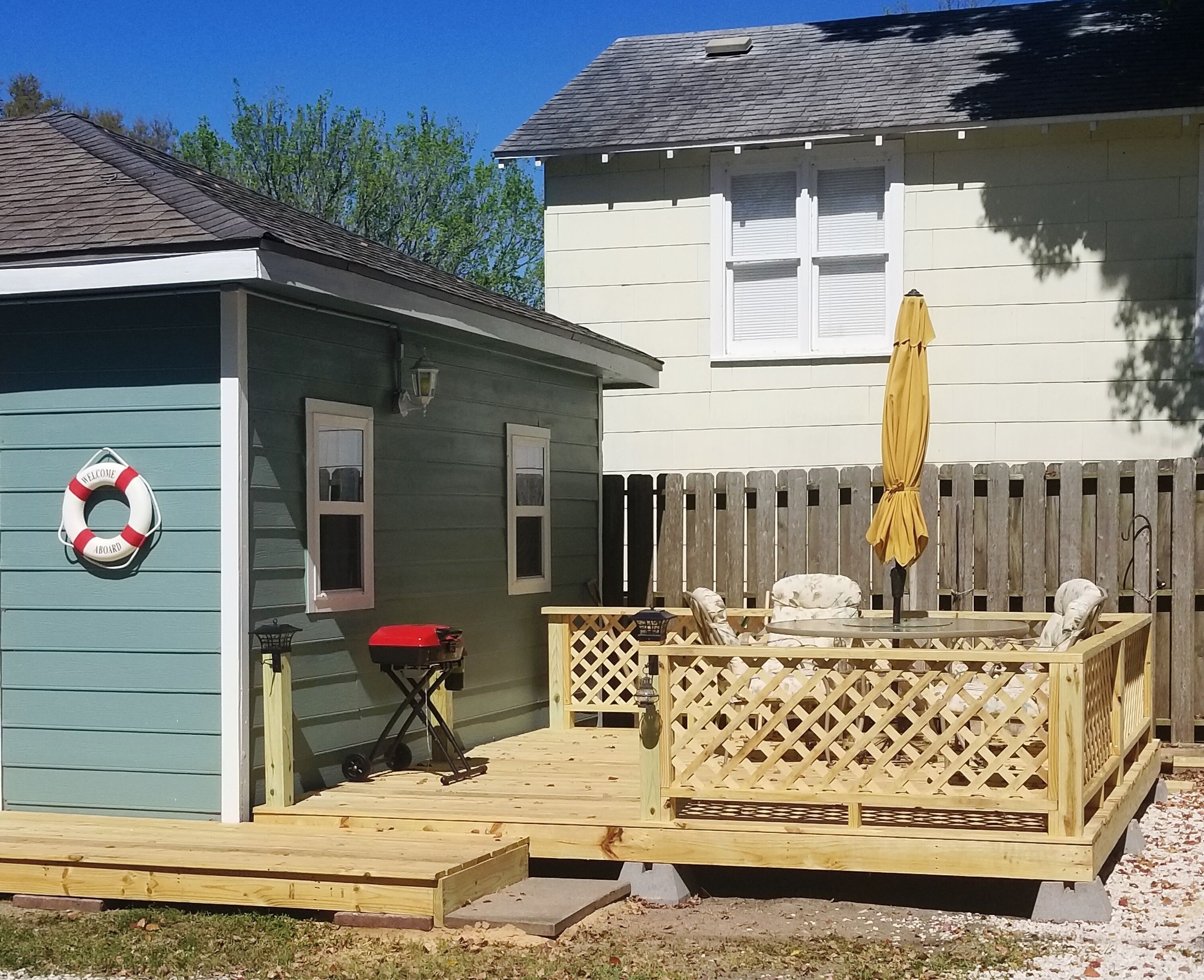 Une petite terrasse en bois, entourée d’une clôture en bois, avec un parasol jaune et une bouée accrochée au mur.