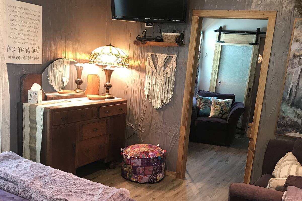 Een knus slaapkamer met een bed, een dressoir, een spiegel, een televisie en een klein rond voetenbankje.