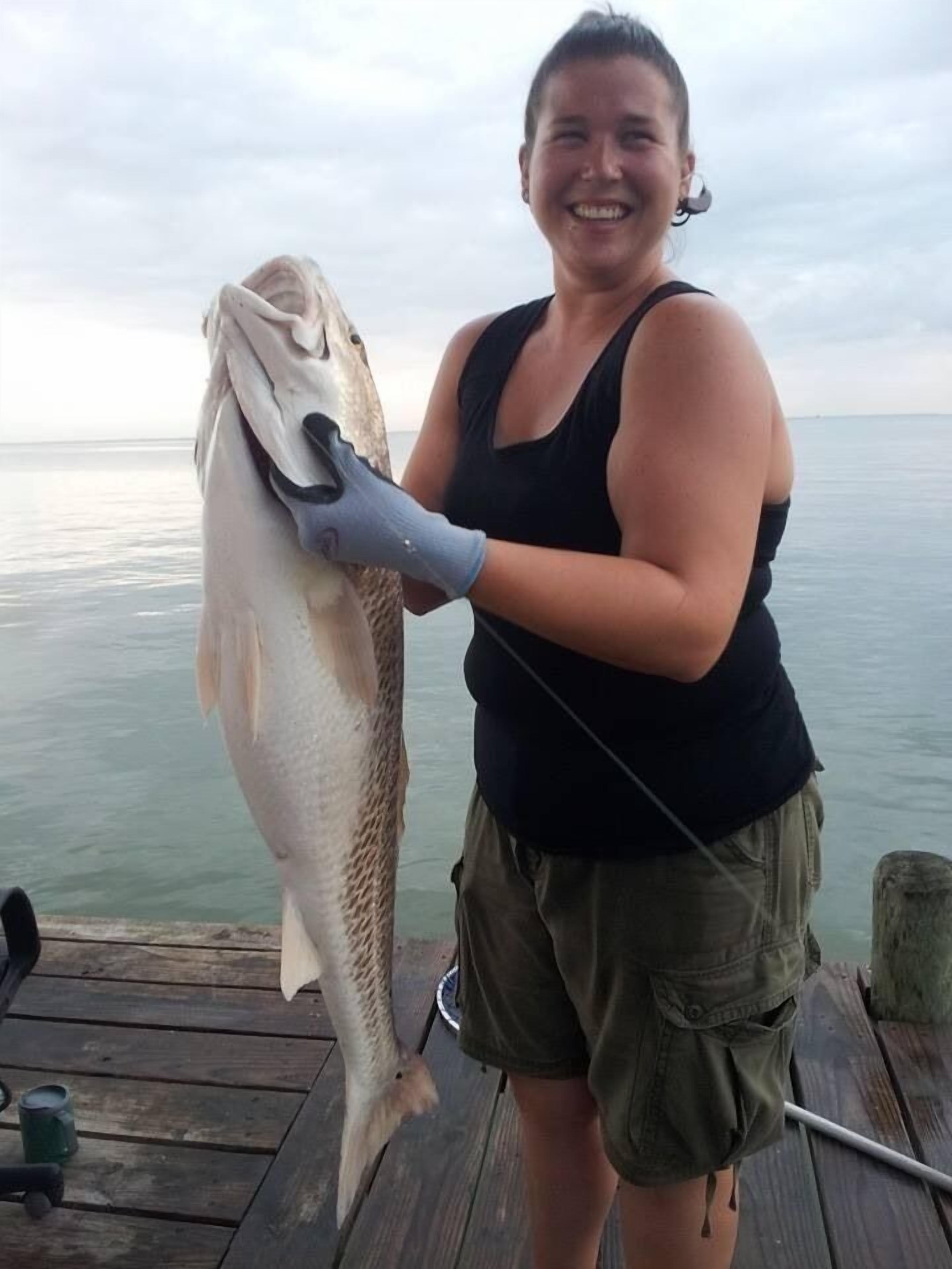 A person holding a large fish on a wooden dock with a calm body of water in the background.