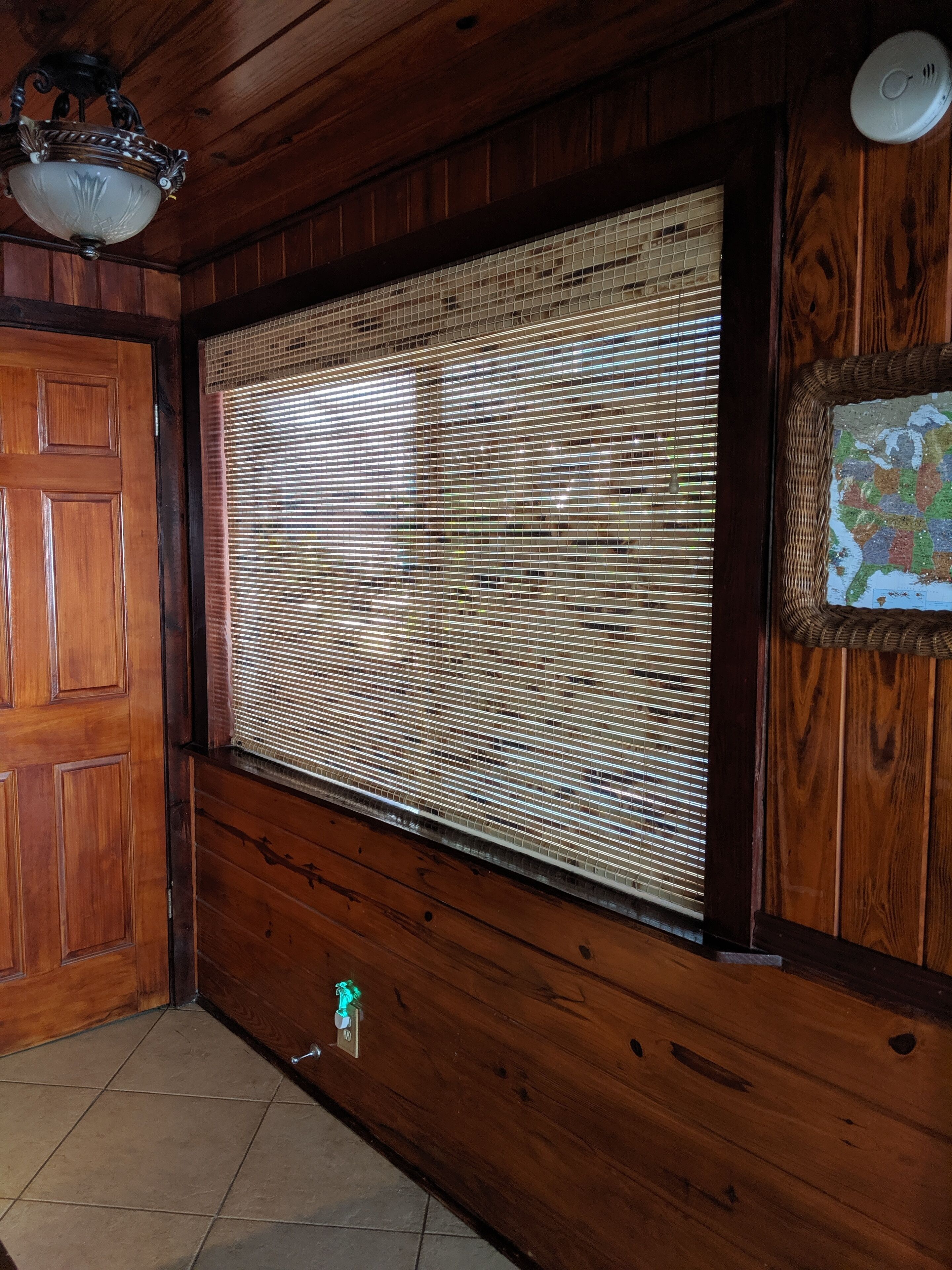 Una habitación con paneles de madera, una ventana grande cubierta por persianas, una puerta y una lámpara fijada a la pared.
