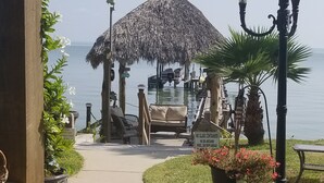 A pathway leading to a thatched-roof structure with a view of the water. There are lounge chairs and a sign indicating a no-glass container policy.