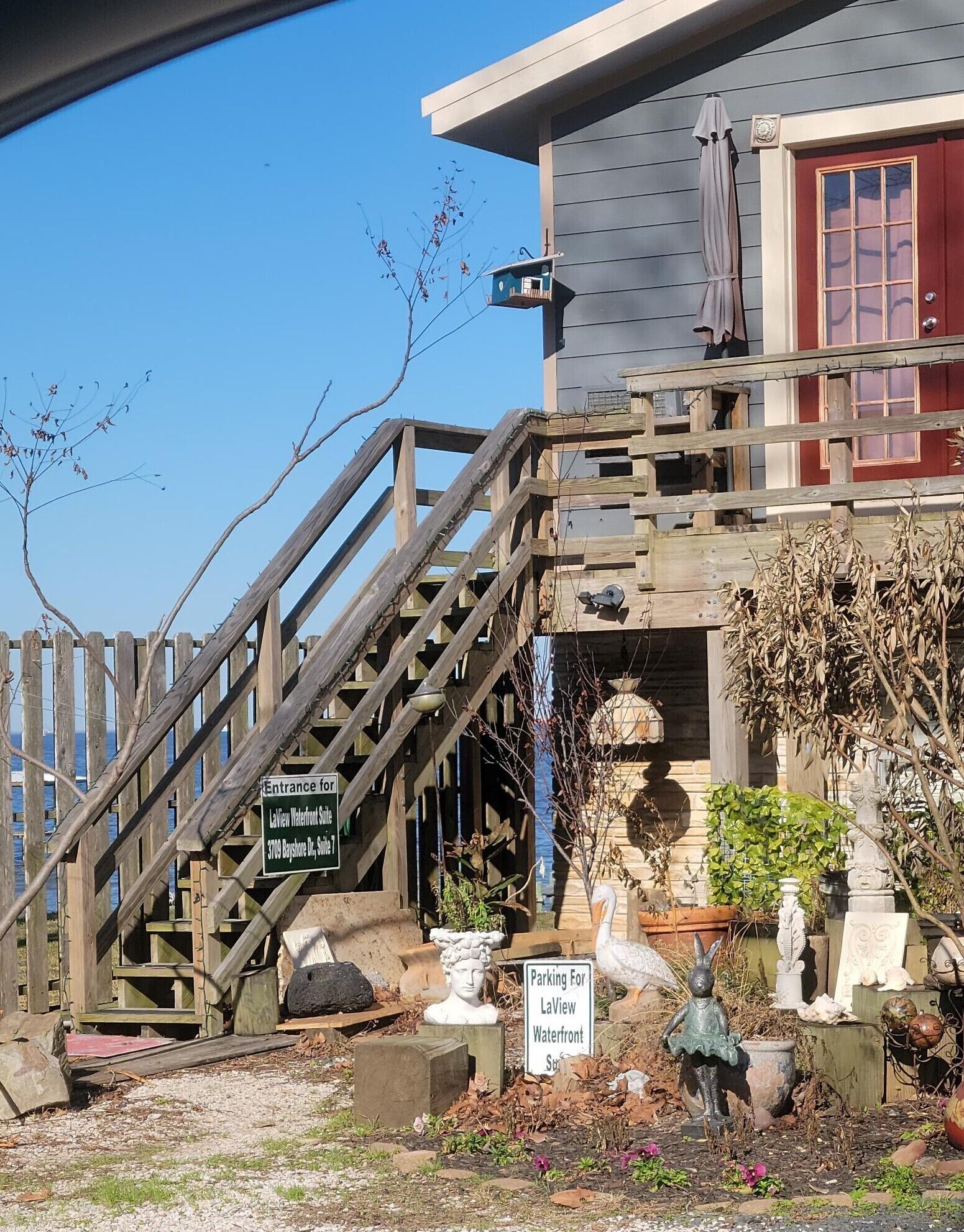 Una escalera de madera lleva a una casa con puerta roja, un cartel de LaFayette Waterfront Suites y diversas estatuas y plantas en el jardín.