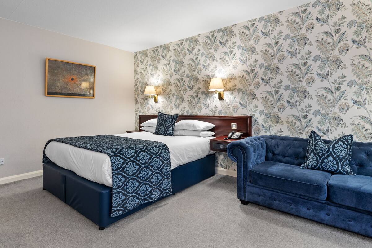Ein Hotelzimmer mit einem Bett, einem blauen Sofa, einem Nachttisch mit Telefon und einer Wand mit floralem Wandpapier.