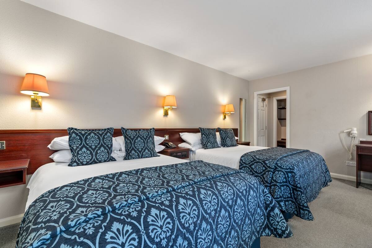 Ein Hotelzimmer mit zwei Betten, jeweils mit blauen, gemusterten Bettdecken, zwei Wandlampen und einem hölzernen Kopfteil.