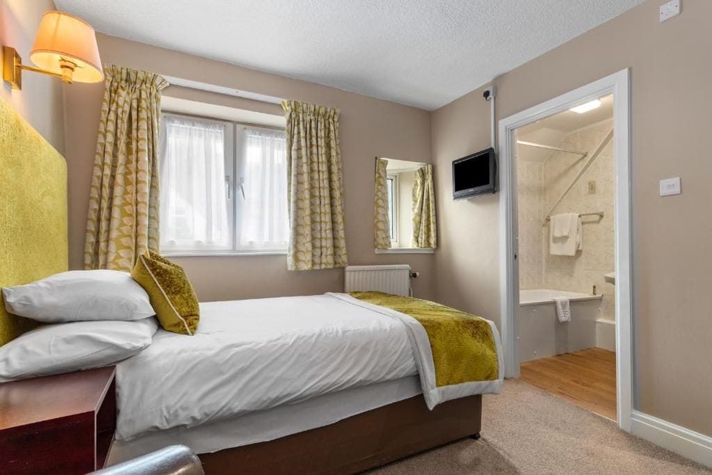A bedroom with a bed, a bedside table, a lamp, a window with curtains, a television, and a bathroom visible through an open door.